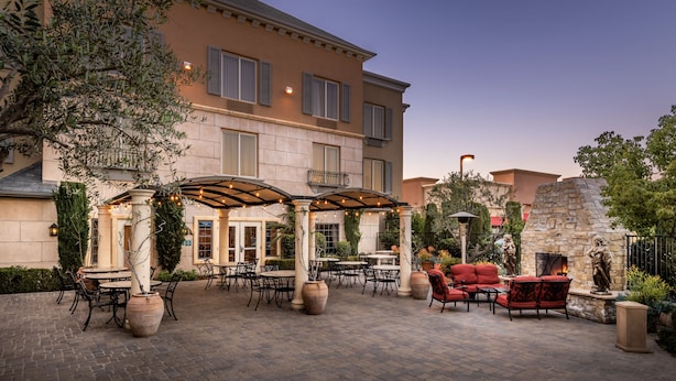 An outdoor patio with tables and chairs, a covered seating area, and a stone wall with a statue. - Ayres Hotel Seal Beach - Cypress (Seal Beach)
