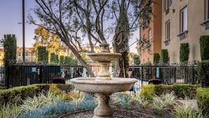 A stone fountain surrounded by a gravel path and various plants, with a fence and buildings in the background. - Ayres Hotel Seal Beach - Cypress (Seal Beach)