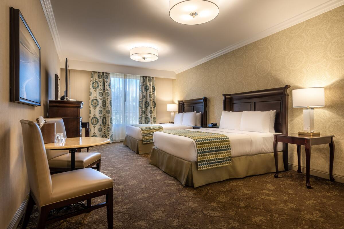 A hotel room with two beds, a desk, a chair, and a lamp.