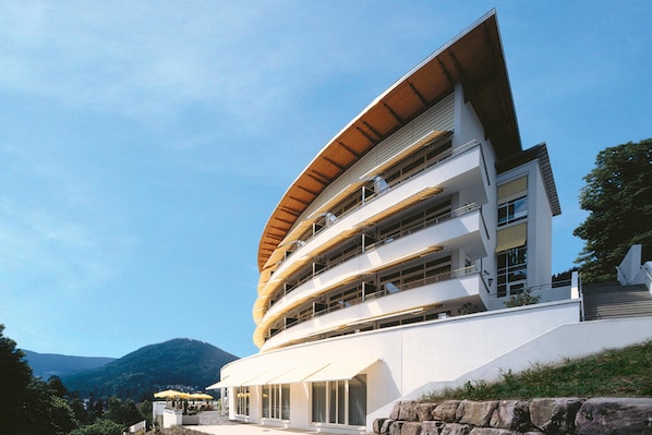 Un edificio moderno de varios pisos con techo curvo, varios balcones y un muro de contención de piedra.
