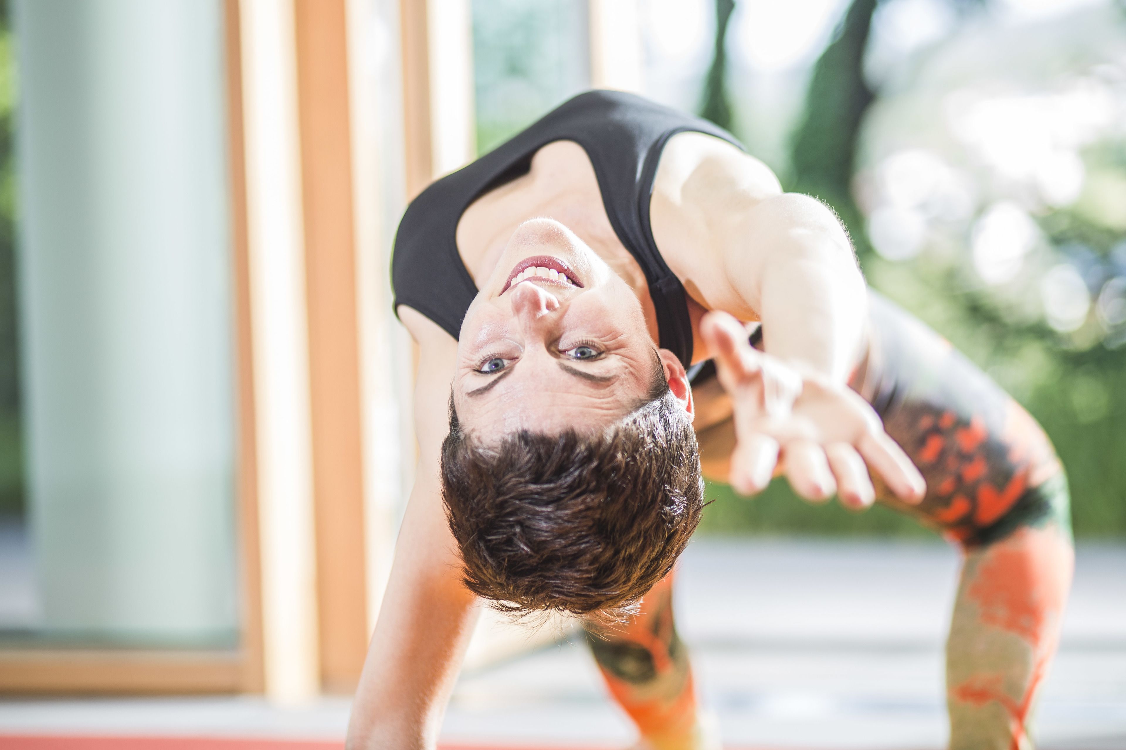 Una persona practicando una postura de yoga, con tatuajes en los brazos, en una habitación con un ventanal grande y suelo rojo.