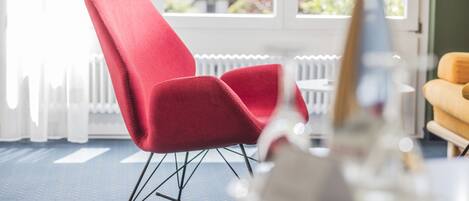 A red chair with a metal frame in a room with a blue carpet, a white radiator, and a window with a view of greenery.
