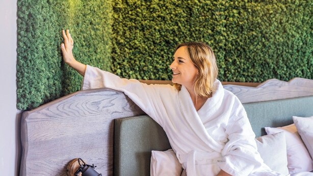 Una persona con bata blanca sentada en una cama, con una pared verde texturizada al fondo.