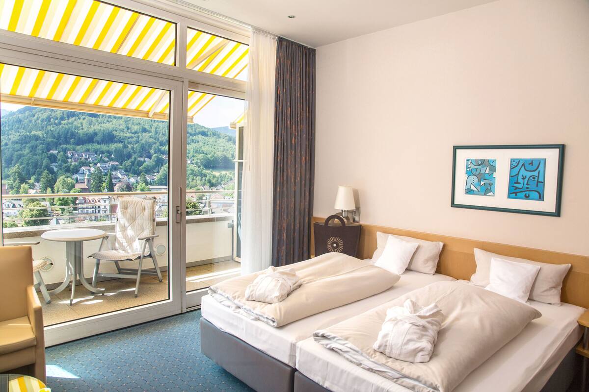 A hotel room with a balcony, a bed with two pillows, a bedside table with a lamp, and a framed picture on the wall.