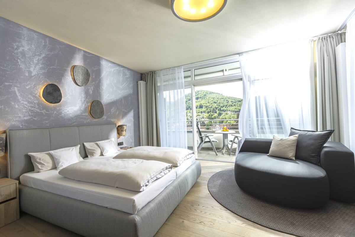 A modern hotel room with a bed, a sofa, and a balcony with a view of the city.