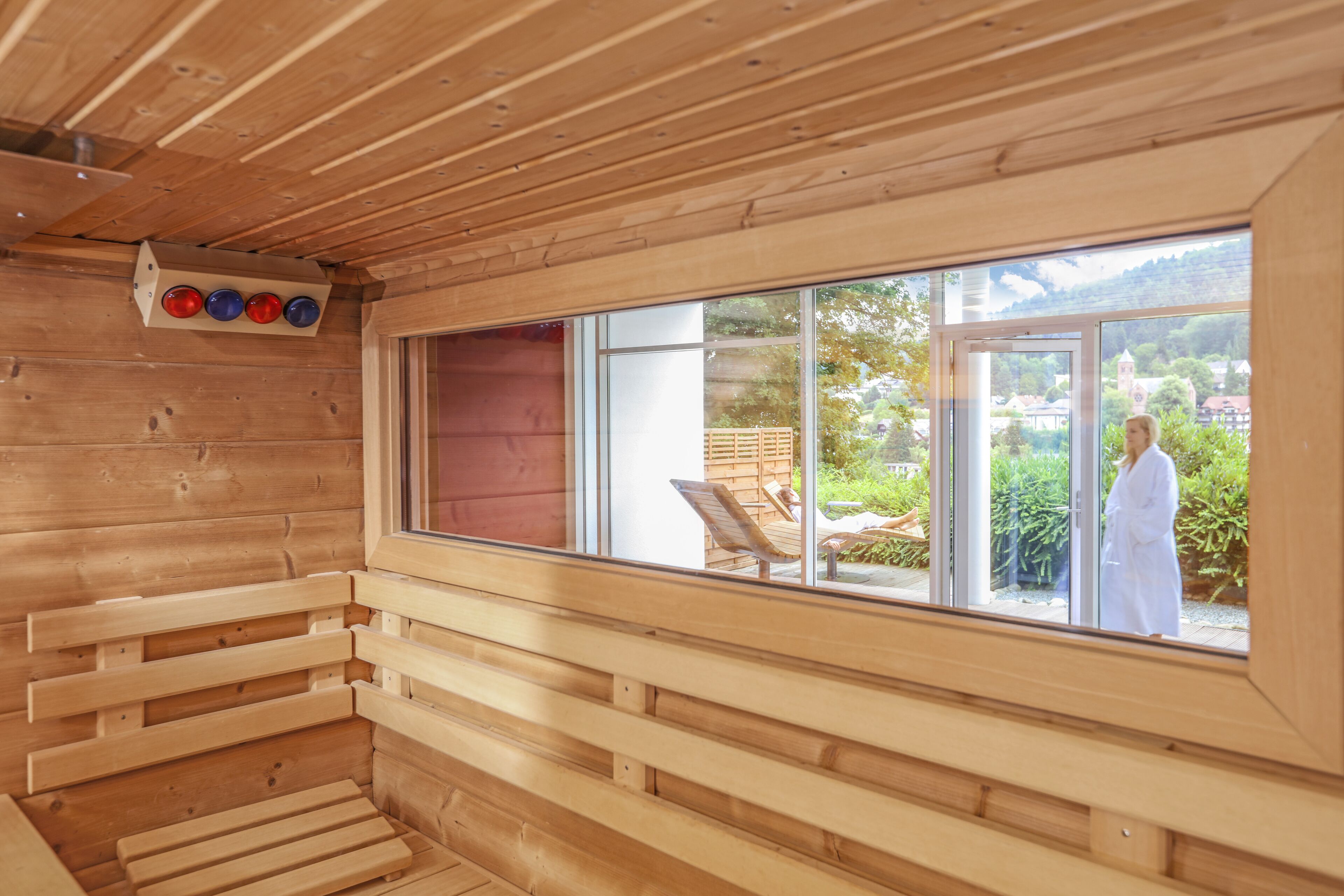 Uma cabana de madeira com sala de sauna, uma janela grande e vista para uma pessoa de roupão branco lá fora.
