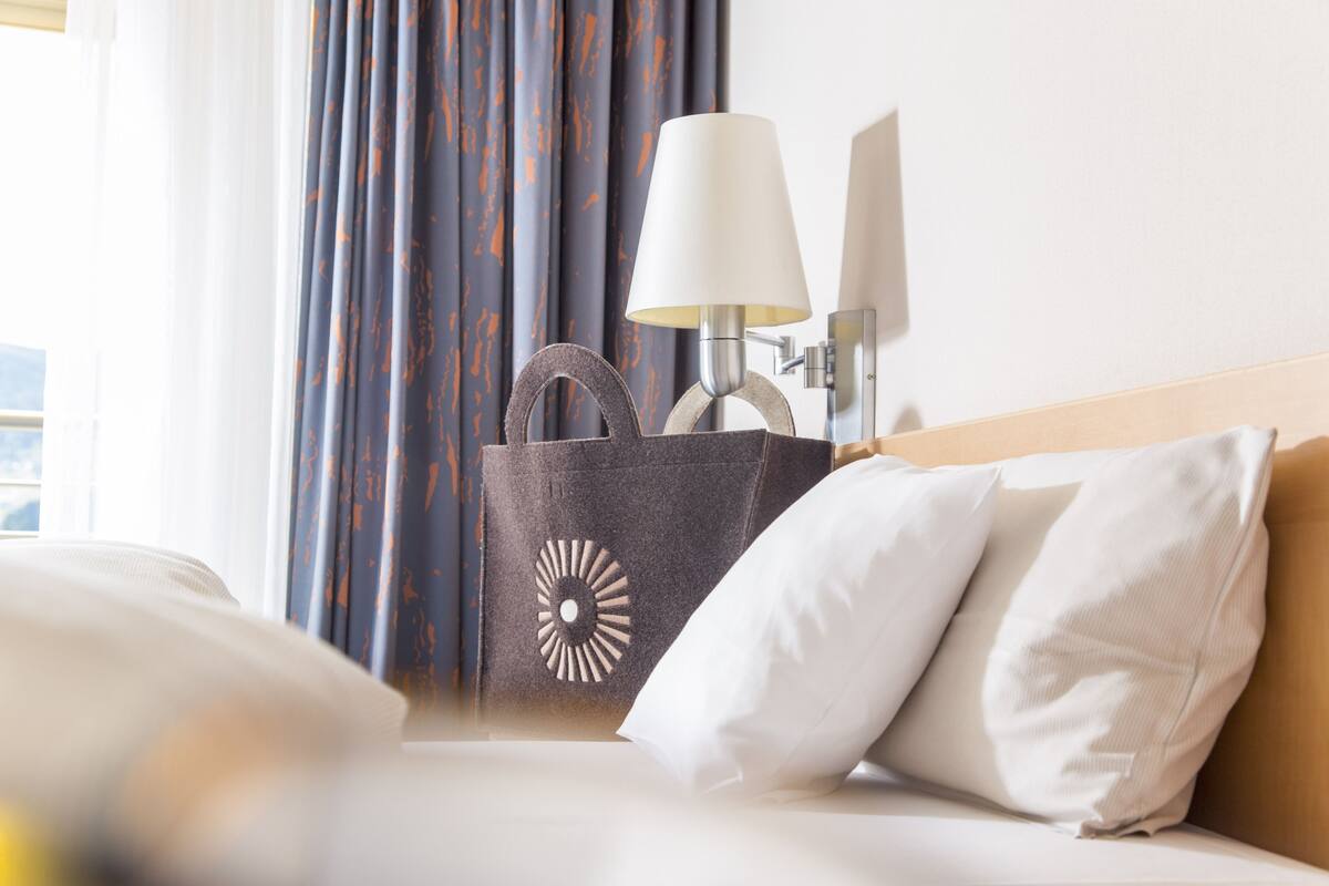A hotel room with a bed, a white pillow, a grey and orange patterned bag, and a lamp.