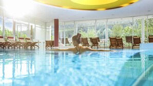 Una piscina interior con paredes de vidrio transparente, sillones de madera y vistas a zonas verdes exteriores.