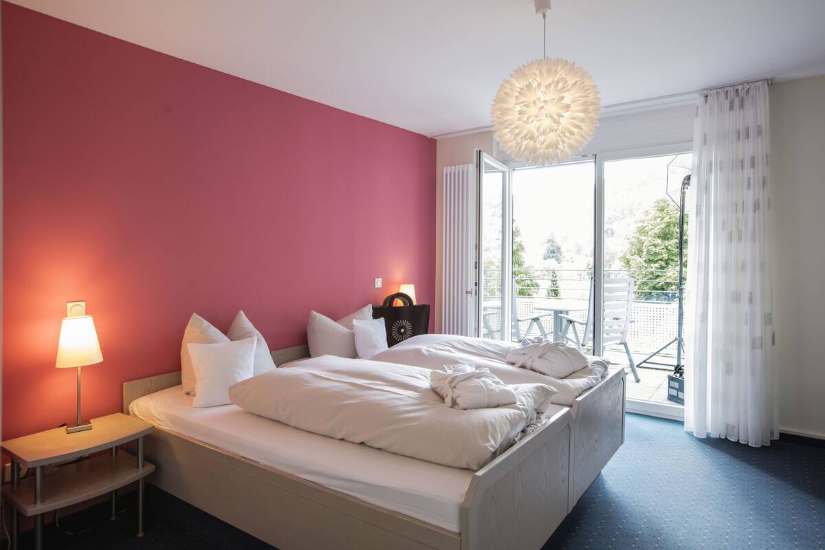 A hotel room with a bed, a nightstand, a lamp, and a view of the outdoors.