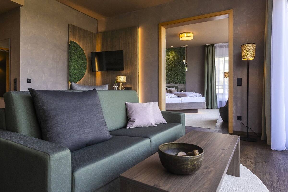 A modern hotel room with a green sofa, a wooden coffee table, a flat-screen TV, and a bed with a green wall and curtains.