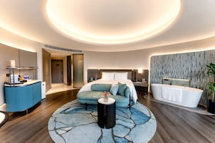 A modern hotel room with a round bed, a sitting area, a bathtub, and a desk with a computer.