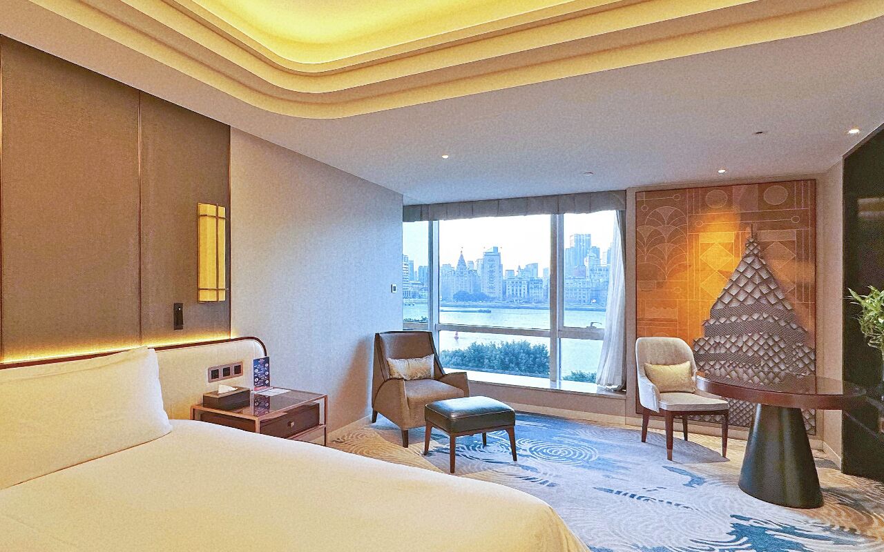 Habitación de hotel con una cama grande, dos sillones, una mesita y vistas a un paisaje urbano a través de amplios ventanales.