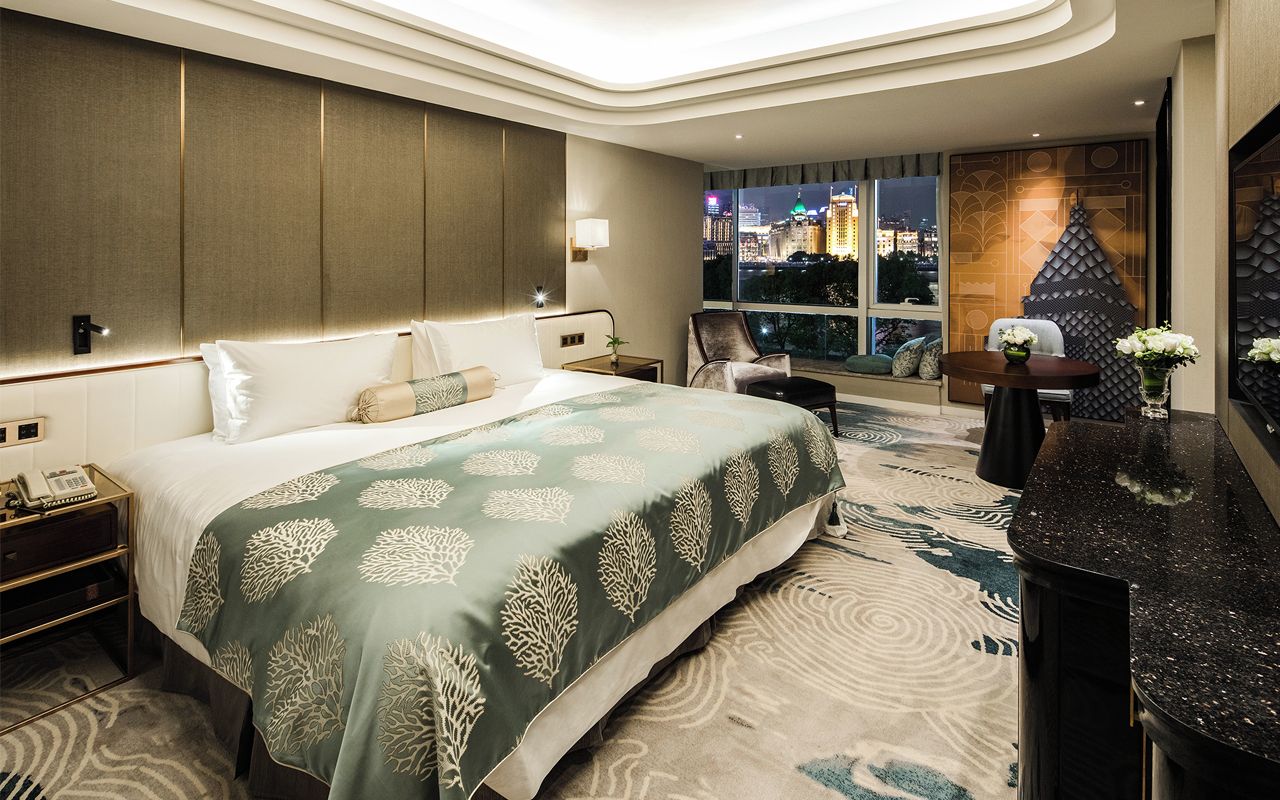 A modern hotel room with a large bed, a nightstand, a desk, and a view of a cityscape at night.