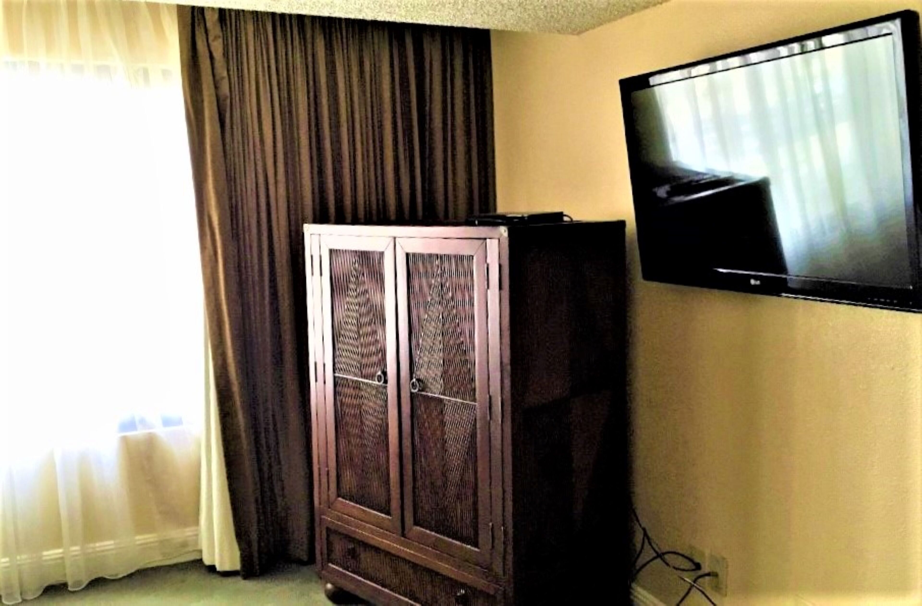 A room with a television mounted on the wall, a dark-colored cabinet, and a window with curtains.