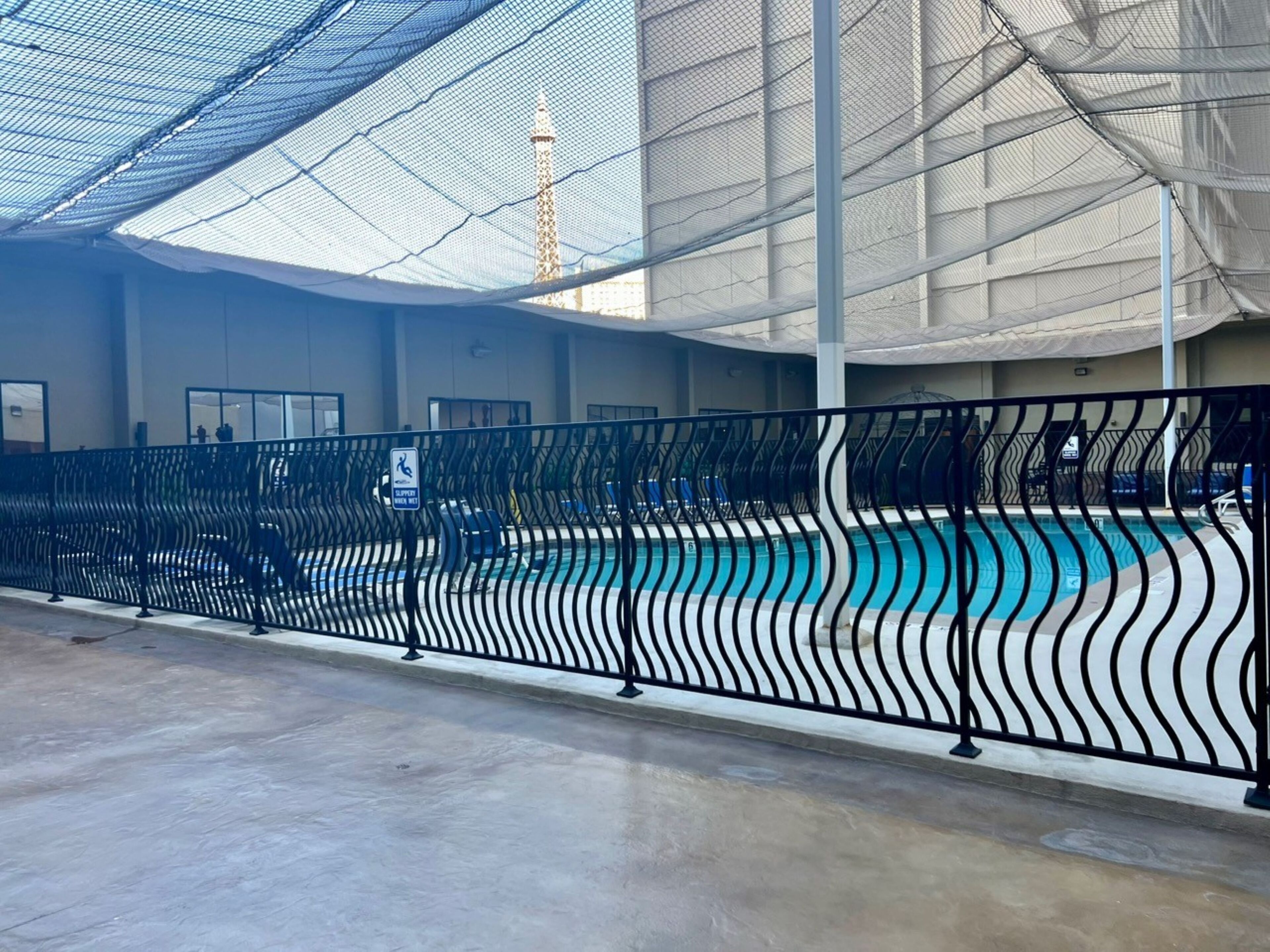 An indoor swimming pool with a black wavy fence, a lifeguard sign, and a view of a tall building in the background.