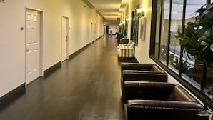 Un pasillo de hotel con una larga galería, puertas a la izquierda y un área de descanso con plantas a la derecha.