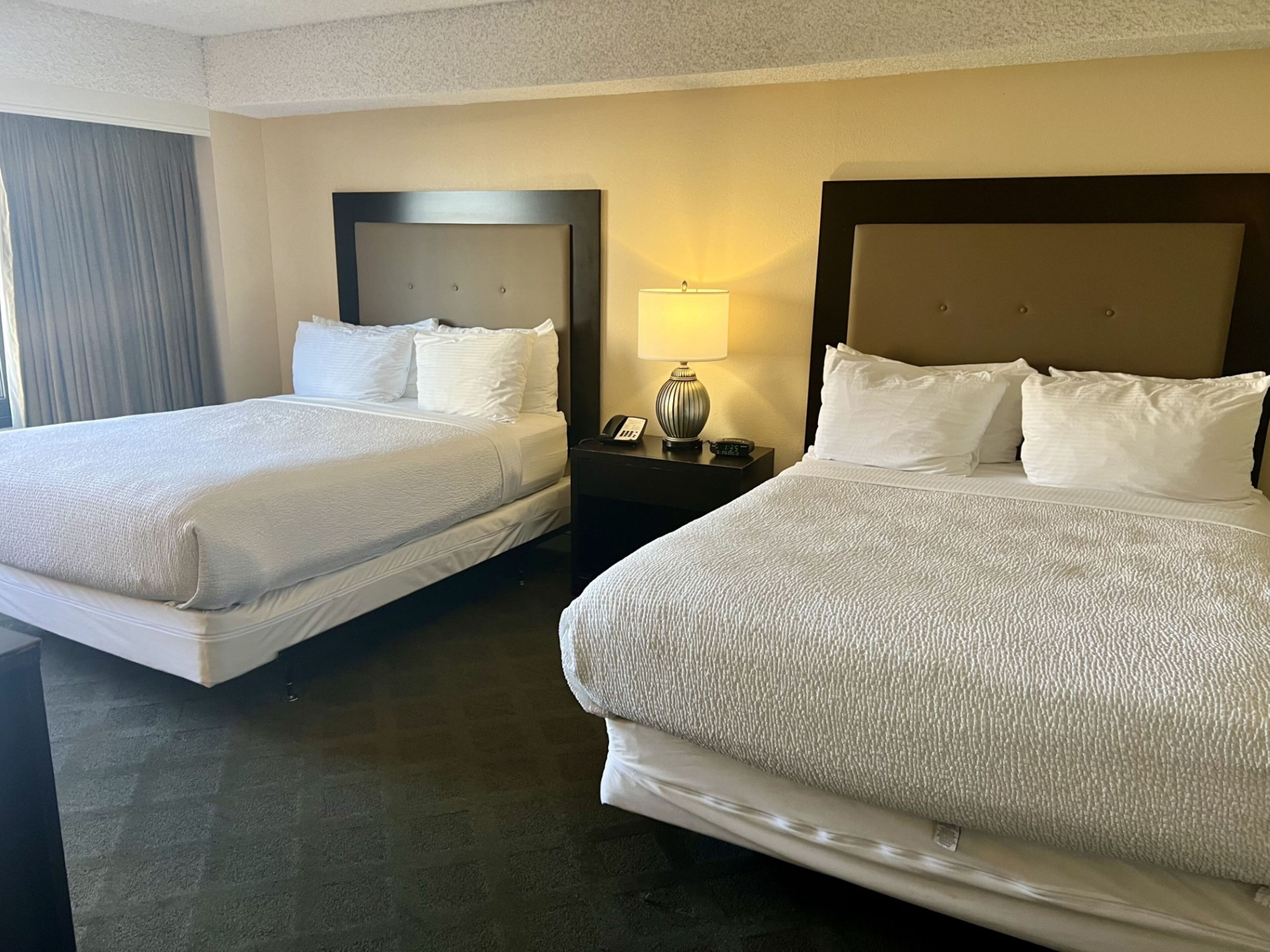 Two twin beds with white linens, a nightstand with a lamp, and a window with curtains.