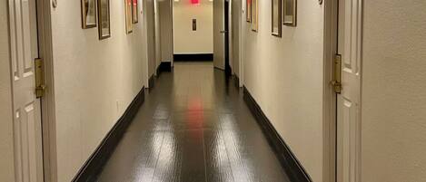 A hotel hallway with doors on both sides, a central elevator, and a red exit sign on the ceiling.