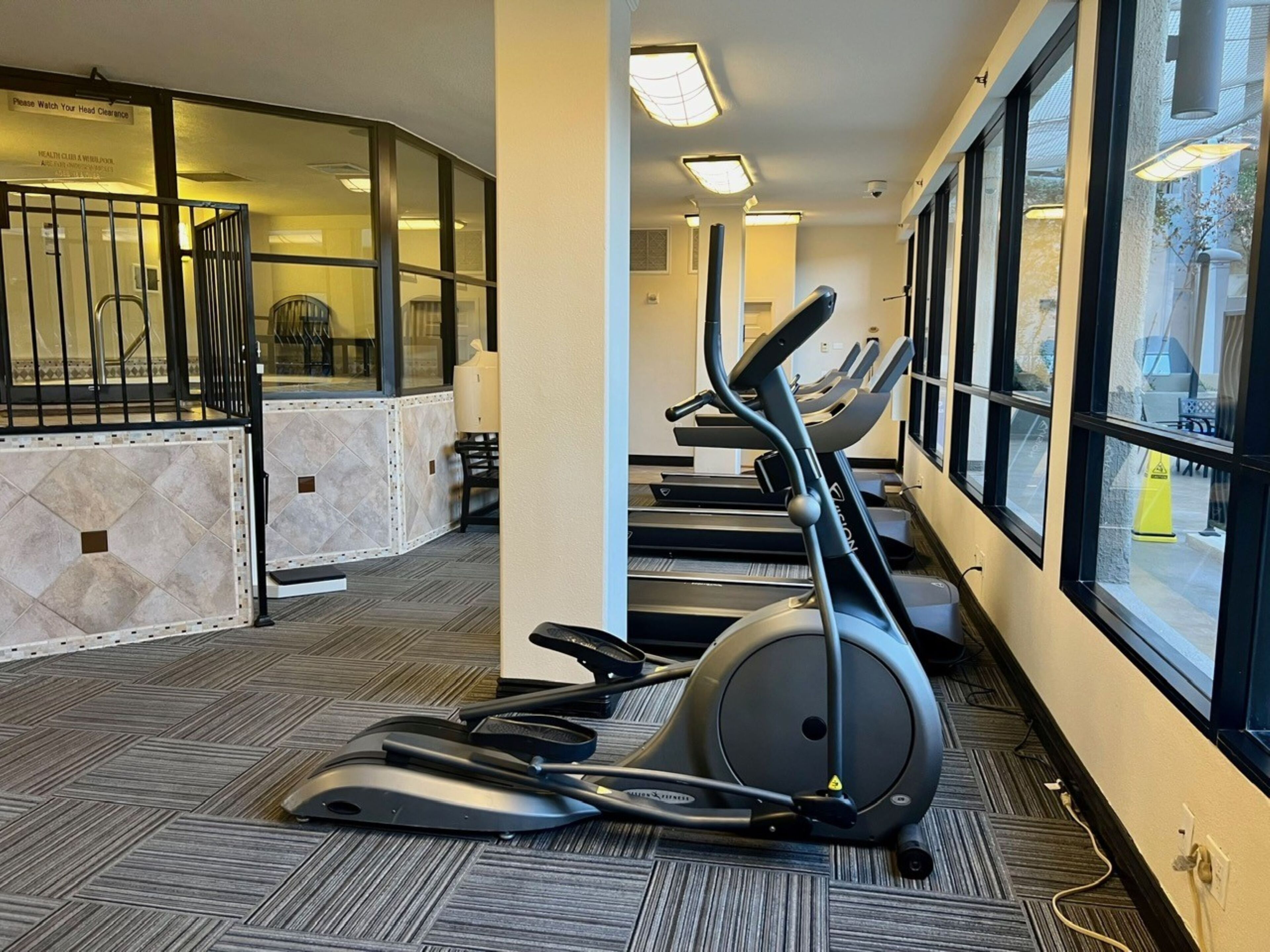 Una palestra con tapis roulant e attrezzature per esercizi, ampie finestre e pavimento piastrellato.