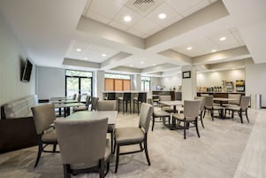 Un comedor moderno con varias mesas y sillas, una zona de estar con sofá, y una cocina equipada con electrodomésticos y un refrigerador.