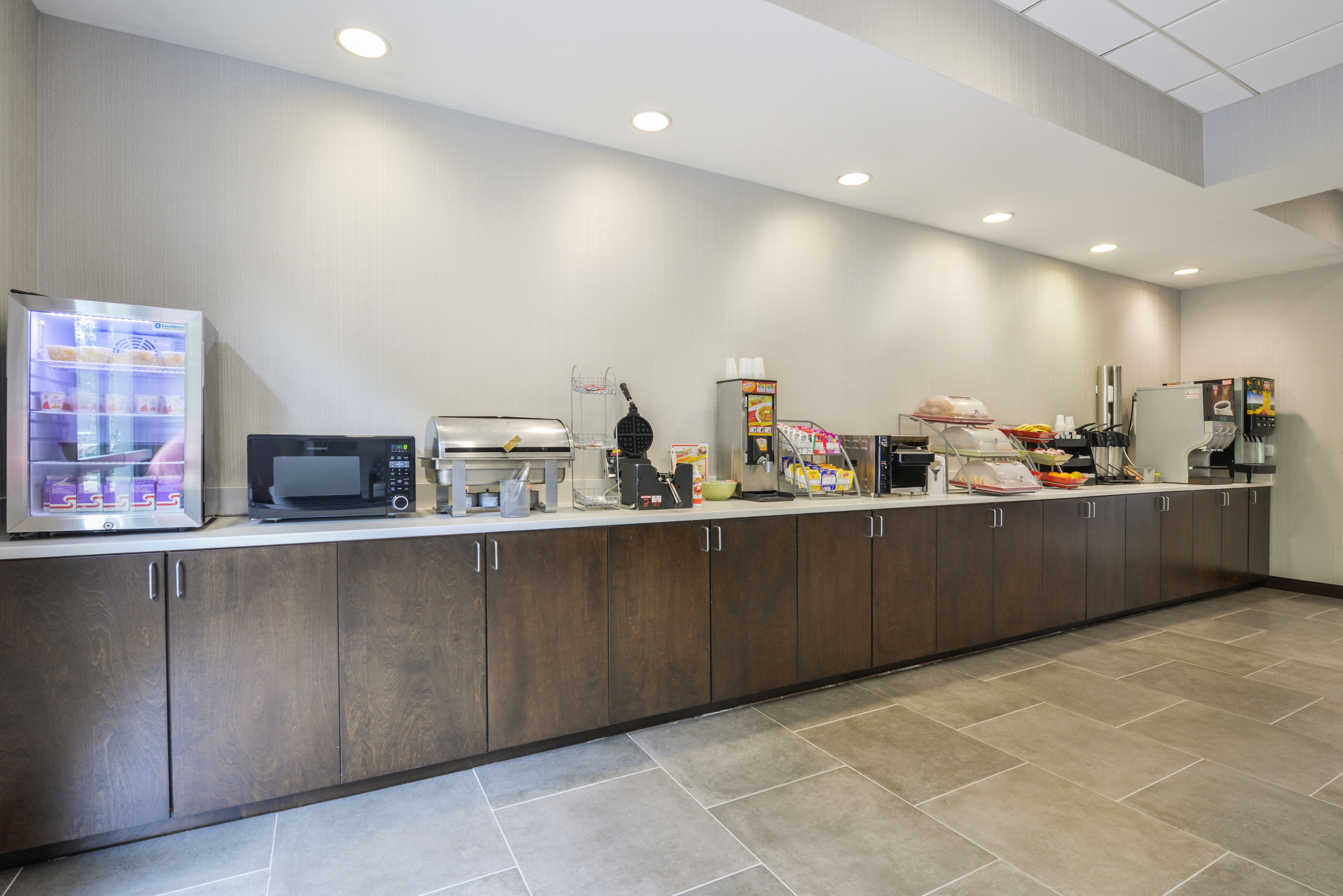 A modern kitchen area with a refrigerator, microwave, coffee maker, and various food items on the counter.