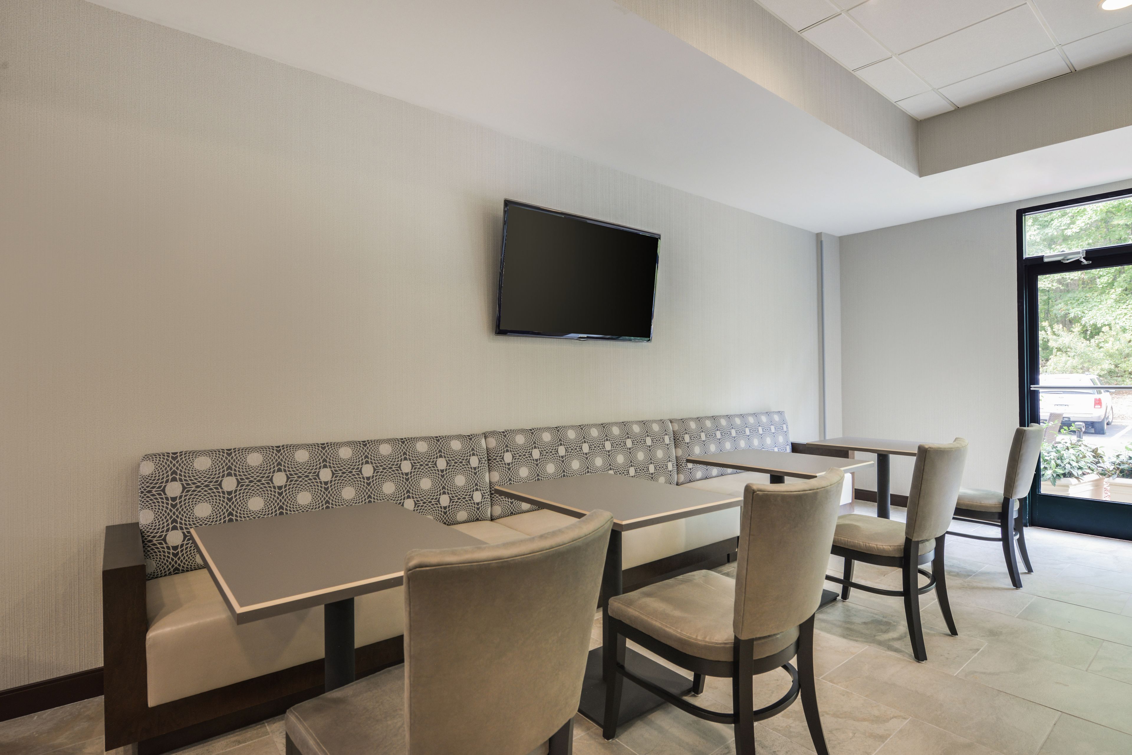 A restaurant interior with a flat-screen TV mounted on the wall, a patterned bench, and several tables with chairs.