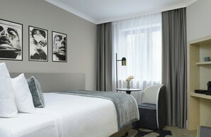 A hotel room with a bed, a chair, a small table with a vase, and a shelf with a cup.