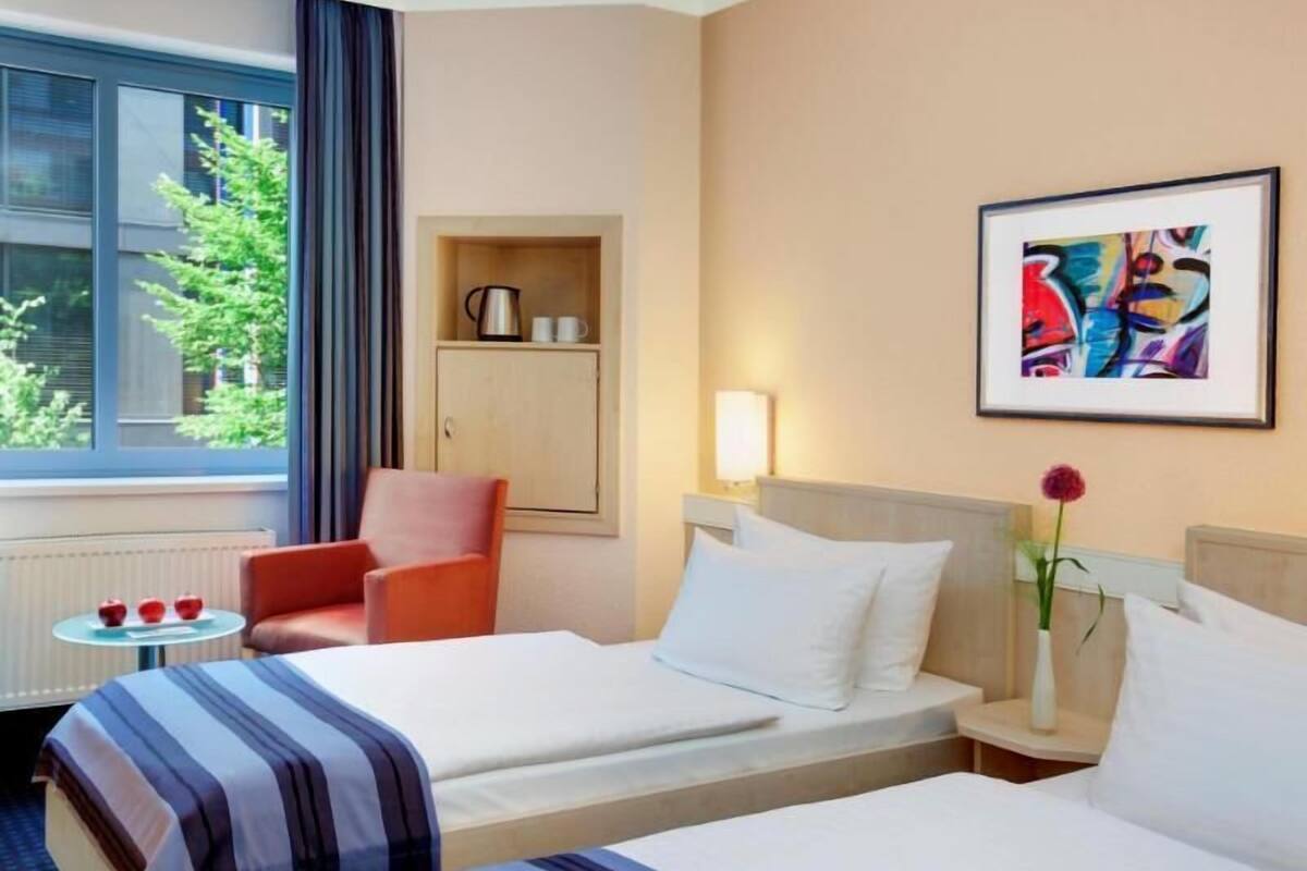 A hotel room with two beds, a small table, a chair, a cabinet, and a window with a view of trees.