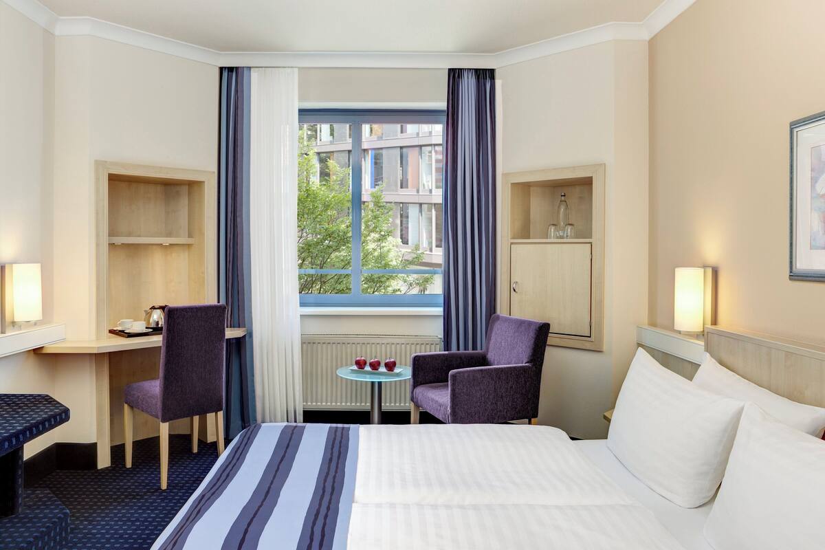 A hotel room with a bed, a desk, a chair, a small table with cups, and a window with curtains.