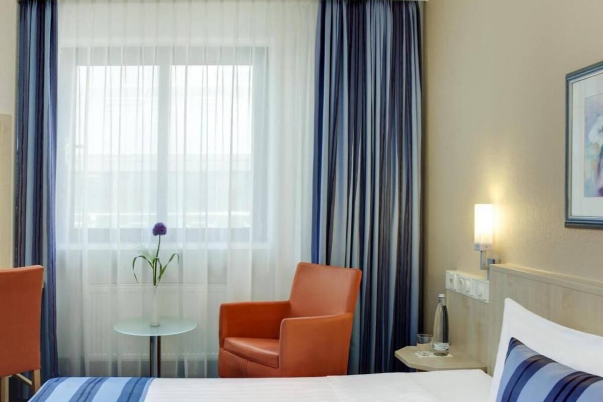 A hotel room with a bed, two chairs, a small table with a vase, a lamp, and a painting on the wall.
