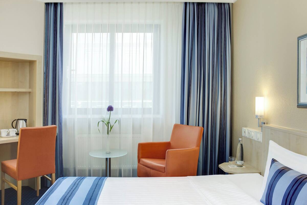A hotel room with a bed, two chairs, a small table with a vase, a nightstand with a lamp, and a window with curtains.