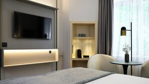 A hotel room with a flat-screen TV mounted on the wall, a bedside table with a lamp, a chair, and a small table with a vase. - Leonardo Hotel Nürnberg City Center (Nuremberg)