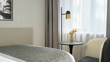 A hotel room with a bed, a chair, a small table with a vase of flowers, and a window with curtains.