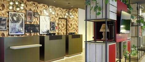 Zona de recepción con pared de madera decorada, varias mesas de trabajo con computadoras y un estante moderno con plantas.