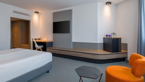 A modern hotel room with a large bed, a desk with a chair, a flat-screen TV, and a seating area with orange chairs.