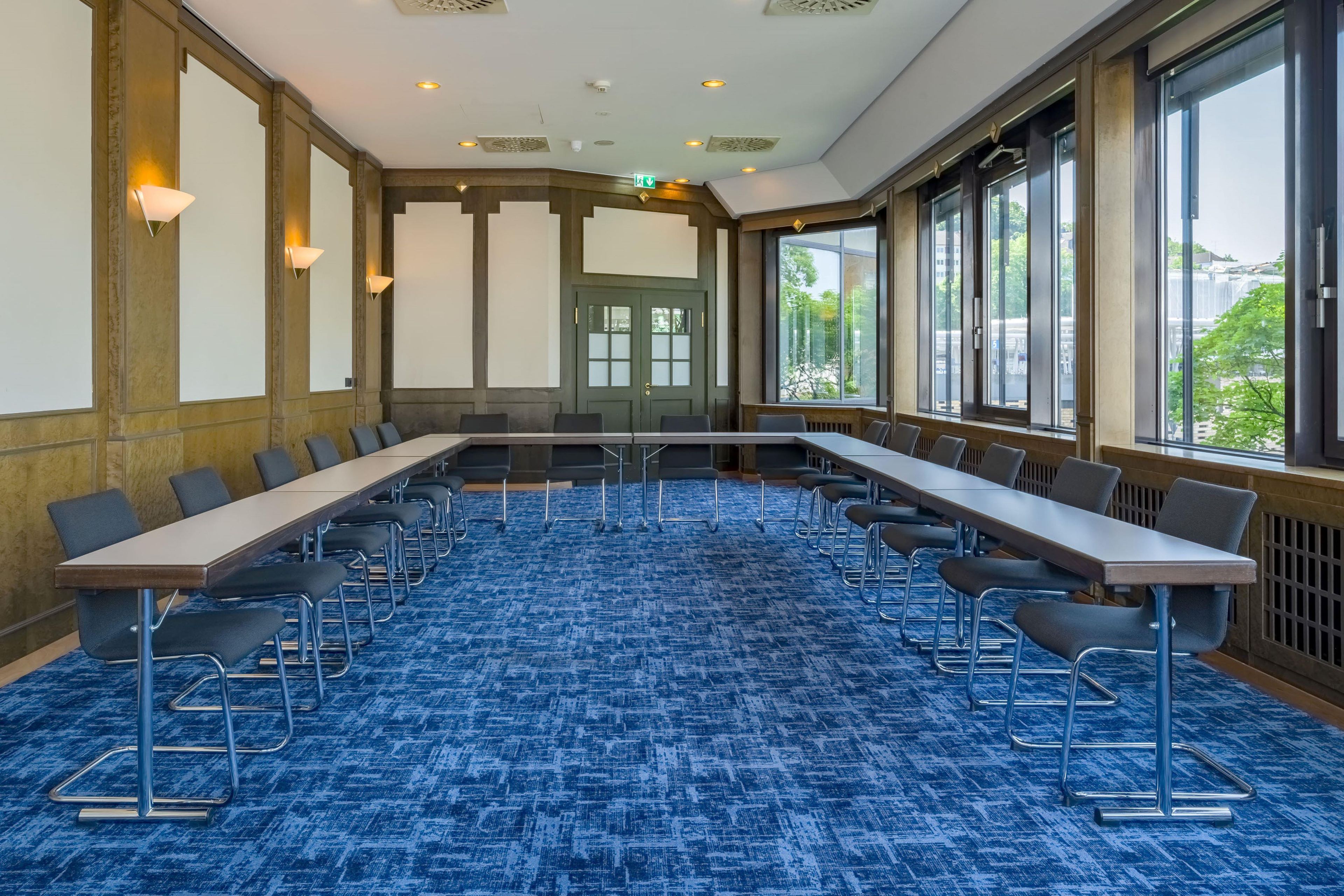 A conference room with a long table and chairs, large windows, and a patterned carpet.
