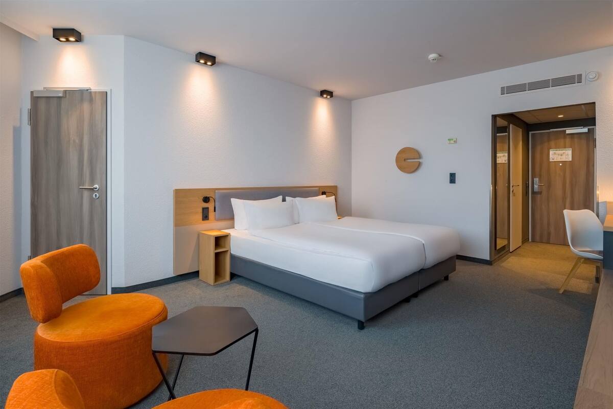A modern hotel room with a large bed, two orange chairs, a small table, and a desk with a chair.