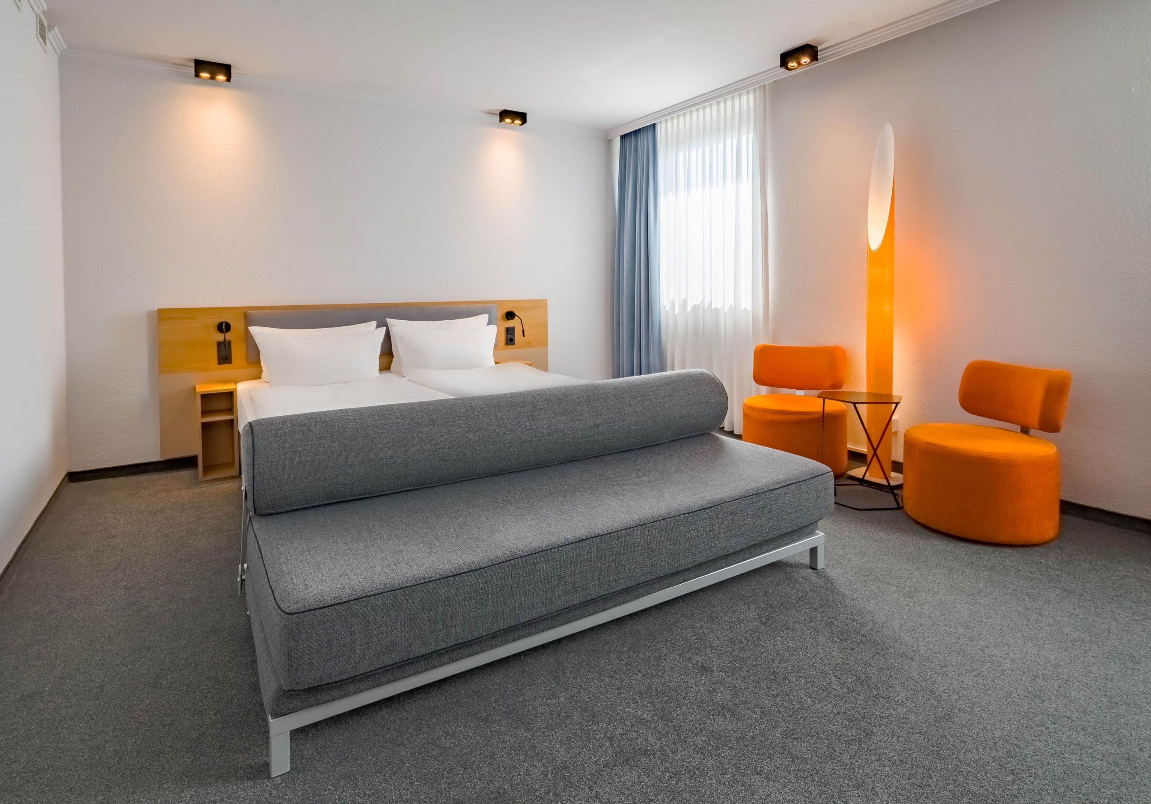 Ein Hotelzimmer mit einem Bett, einer Couch, zwei Sesseln, einer Lampe und einem fenster mit Vorhängen.