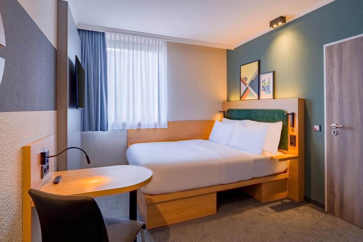 A compact hotel room with a bed, a desk, a chair, a TV, and a door.