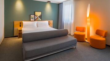Ein Hotelzimmer mit einem Bett, einer Couch, zwei Sesseln, einem Nachttisch, einer Lampe und einem fenster mit Vorhängen.