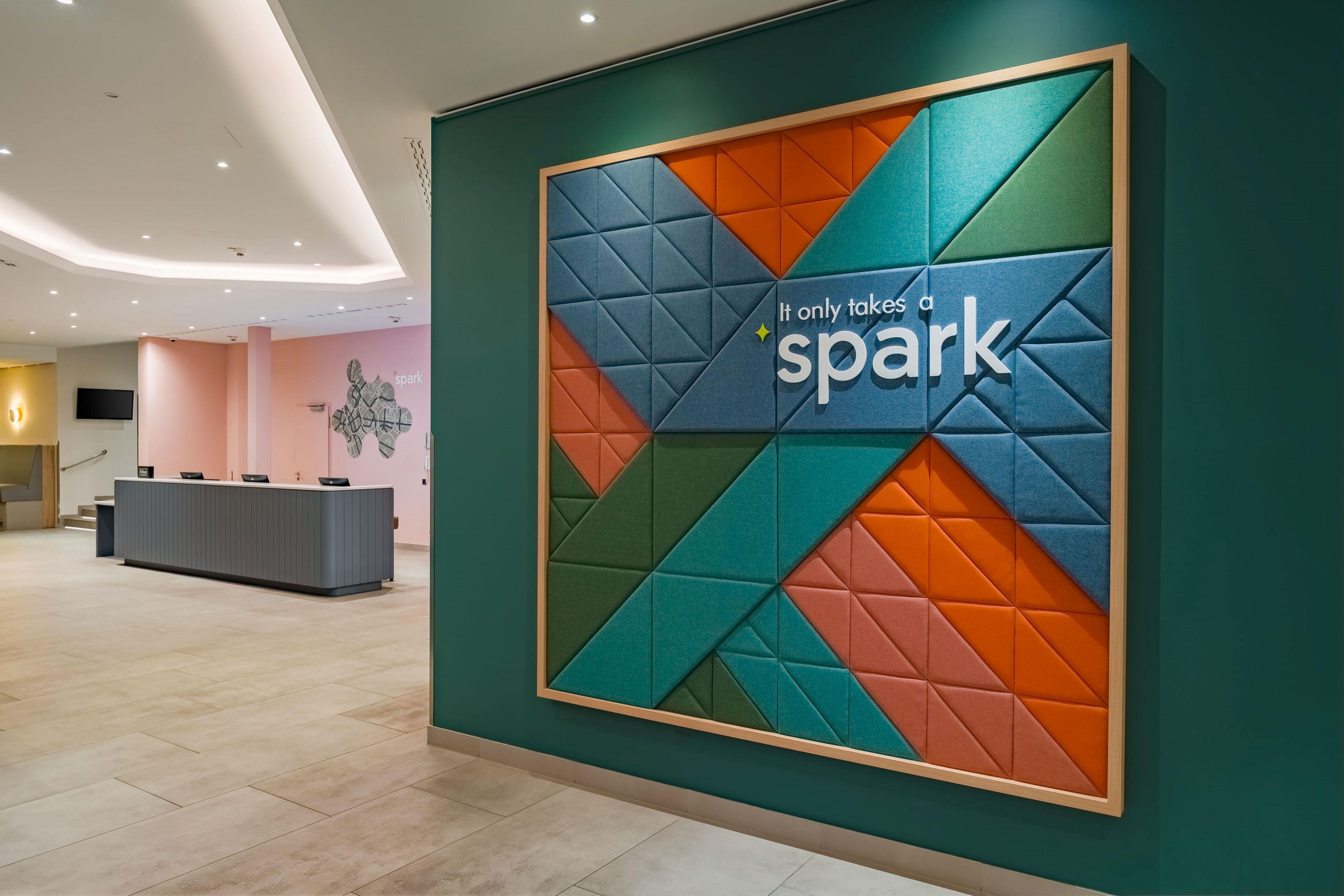 Ein Empfangsbereich mit modernem Design, einer Rezeption und einer Wand mit geometrischen Mustern und dem Text 'It only takes a spark'.