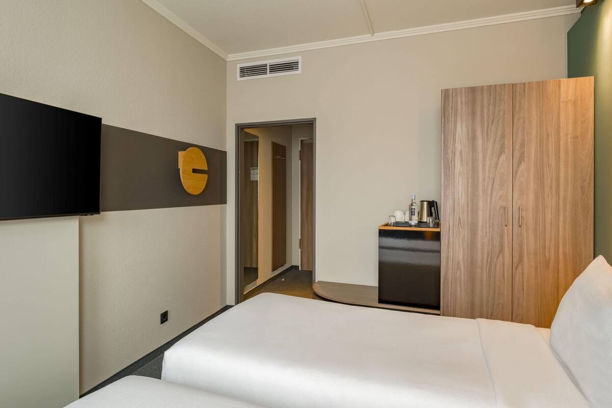 A hotel room with a bed, a wooden wardrobe, a small kitchenette, and a wall-mounted TV.
