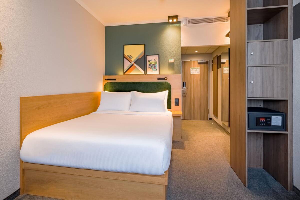 A hotel room with a bed, a wooden headboard, a wardrobe, and a wall with artwork.