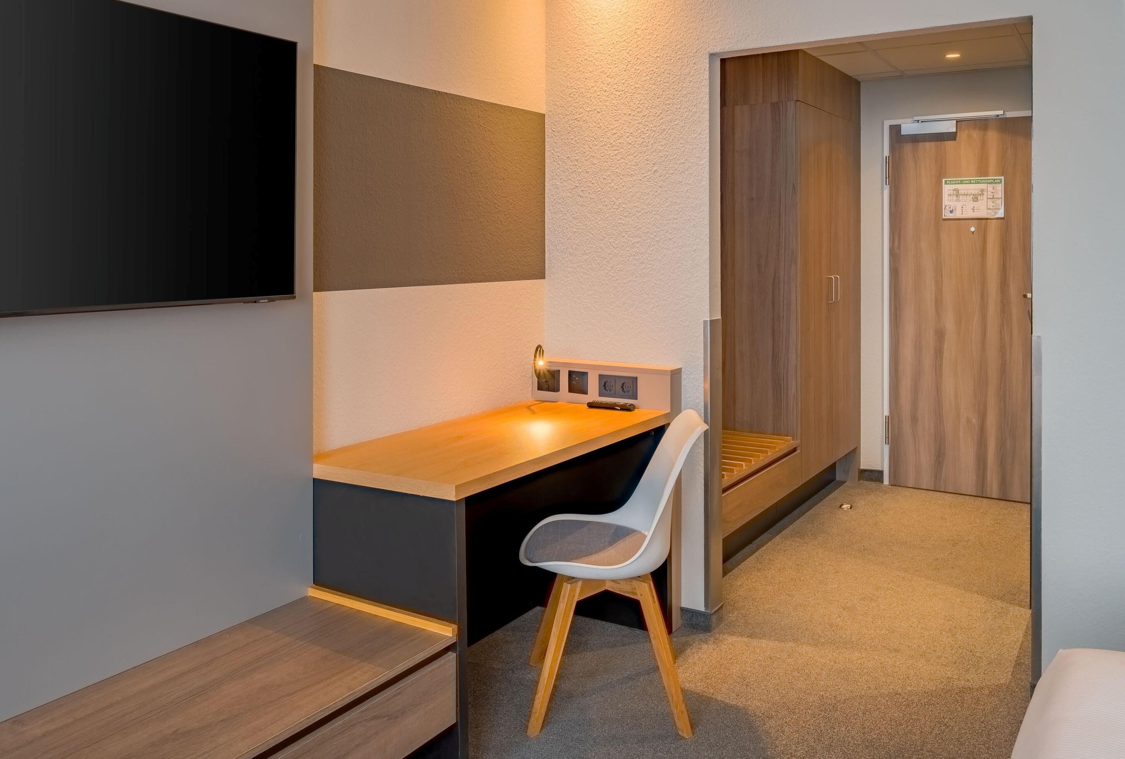 Una habitación de hotel moderna con un escritorio de madera, una silla blanca, un televisor de pantalla plana montado en la pared y un armario de madera.