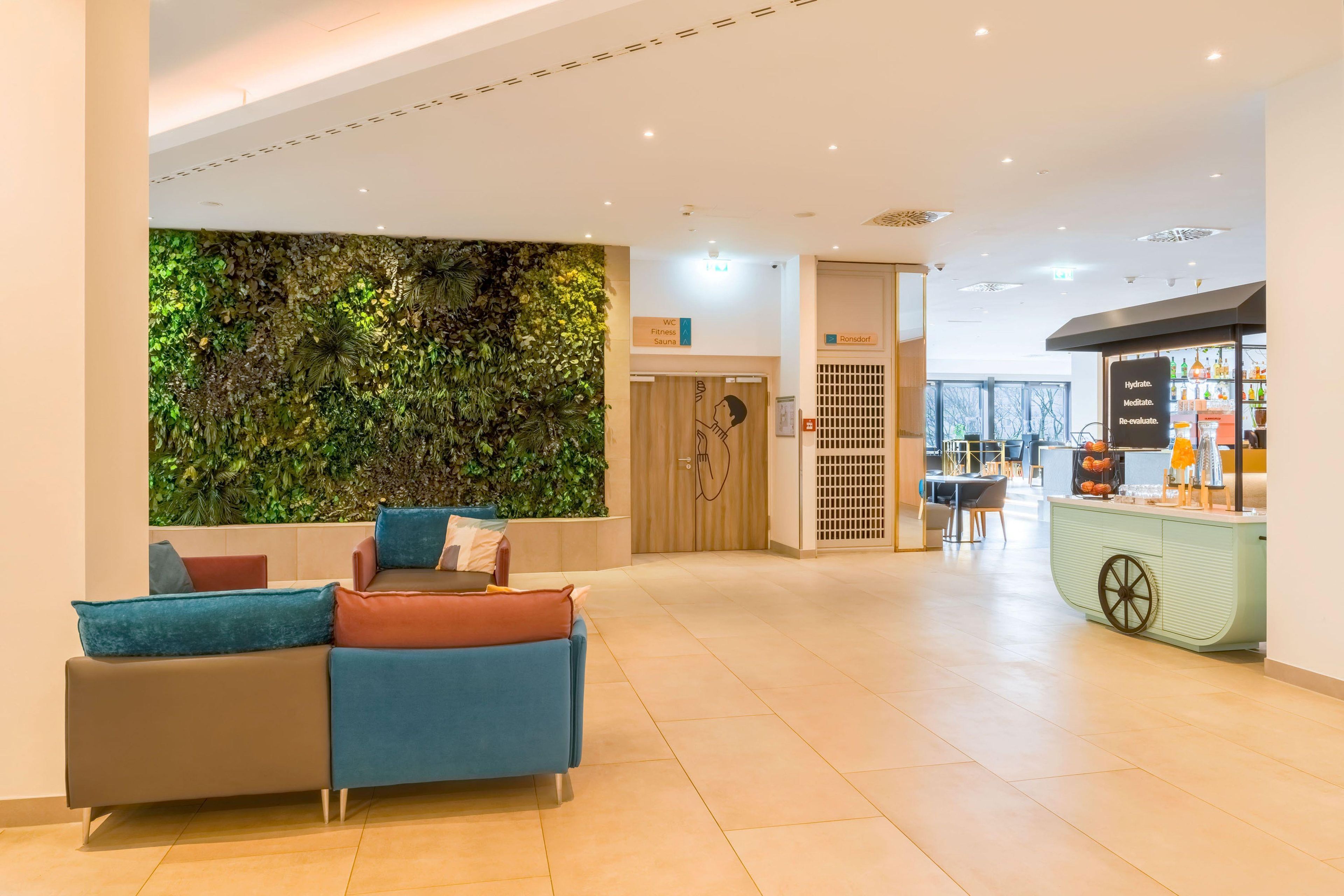 Eine Lobby mit einer Rezeption, einer Sitzecke mit Sofas, einer begrünten Wand und einer Erfrischungsbar.
