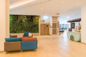 A lobby with a reception desk, a seating area with sofas, a wall with greenery, and a refreshment bar.