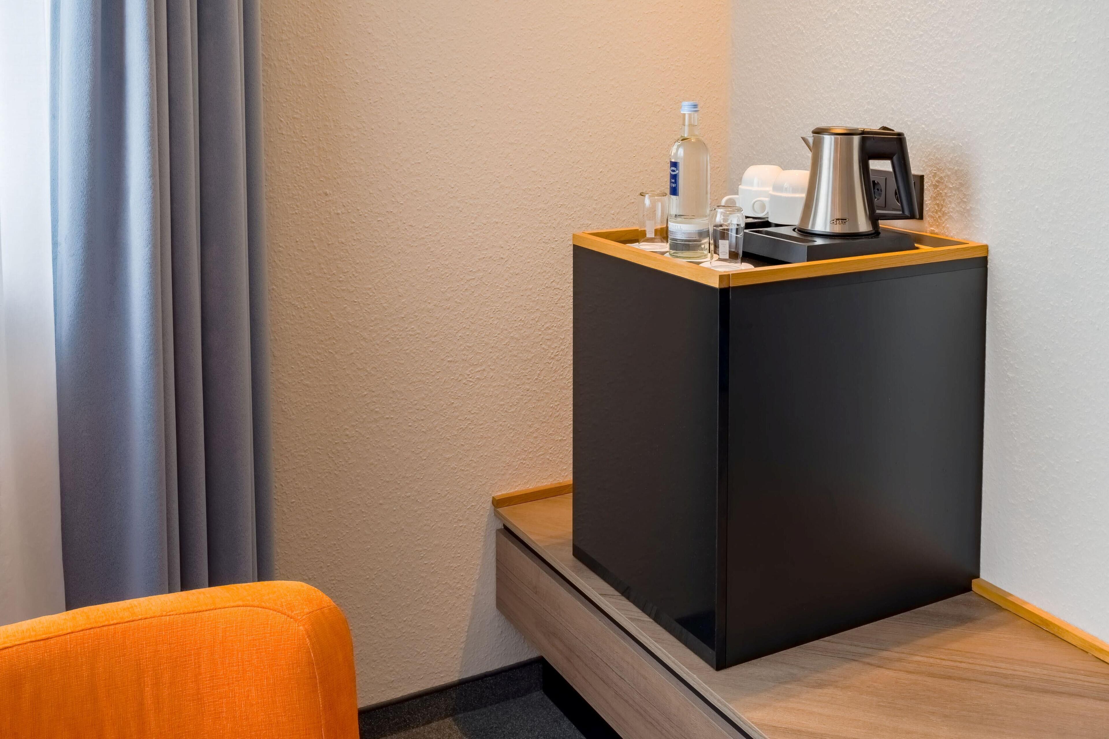 Une chambre d’hôtel avec une table d’appoint en bois, un meuble noir contenant une bouilloire, une bouteille et des verres.