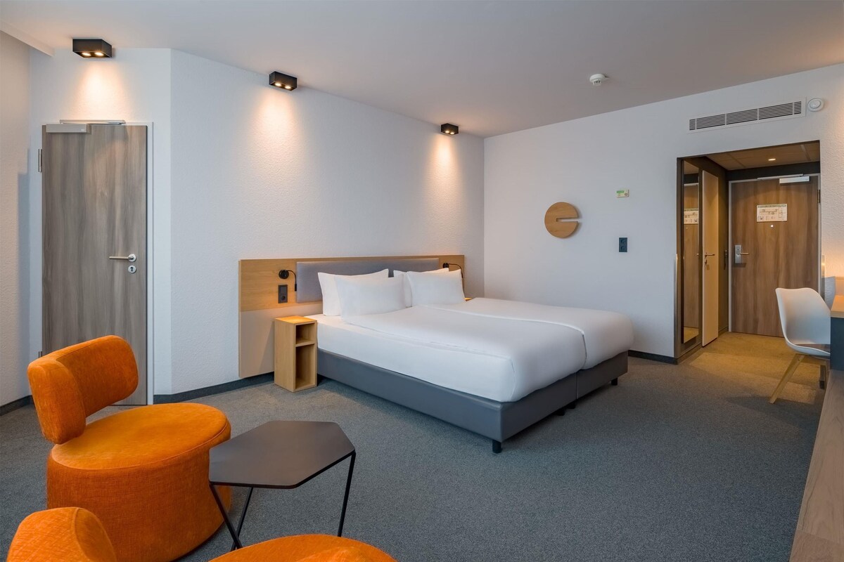 Ein modernes Hotelzimmer mit einem großen Bett, zwei orangefarbenen Sesseln, einem kleinen Tisch und einem Schreibtisch mit Stuhl.