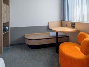 A compact hotel room with a bed, a small table, a chair, a bedside table, a wall-mounted control panel, and a window with curtains. - Spark by Hilton Wuppertal City Centre (Wuppertal)
