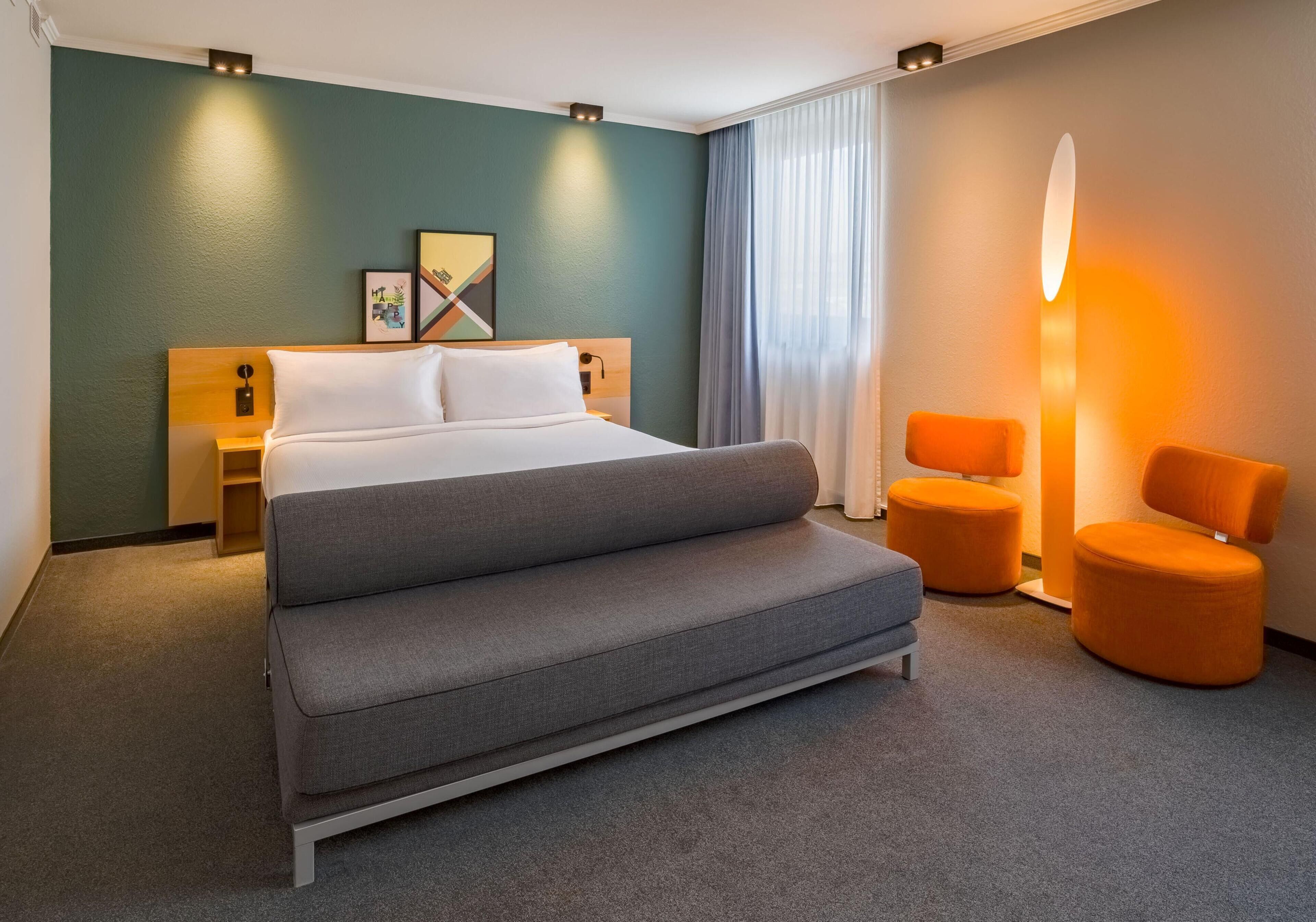 A modern hotel room with a large bed, a sofa, two orange chairs, a nightstand, and a wall-mounted lamp.